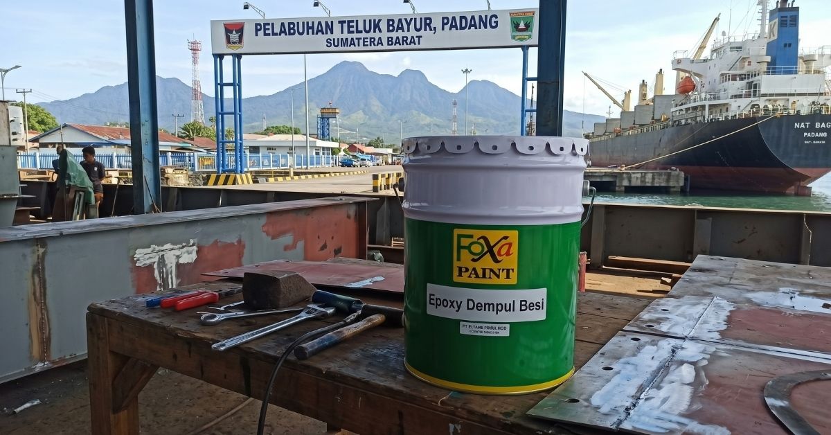 Kaleng Foxapaint Epoxy Dempul Besi dan peralatan kerja di atas meja dengan latar belakang gerbang Pelabuhan Teluk Bayur Padang, kapal kargo, dan pegunungan.